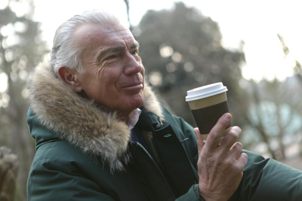 Elderly man enjoying coffee in the winter park wearing a fur-lined coat.