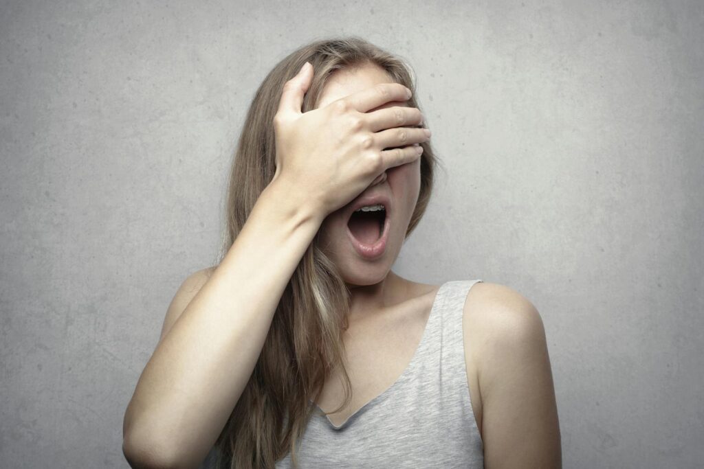 A surprised woman covers her face with one hand, expressing shock and amazement against a gray background.