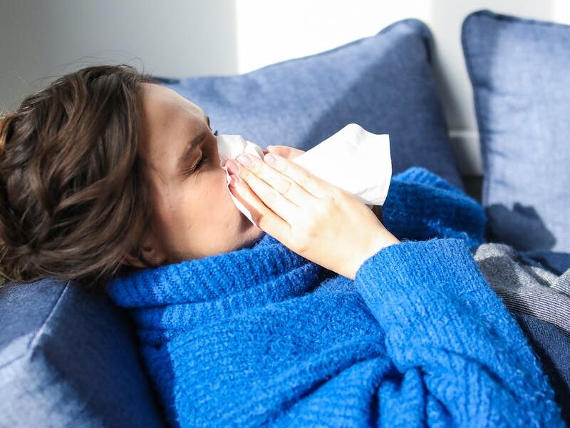 A woman in a blue sweater uses a tissue, lying indoors, suggesting illness or flu symptoms.