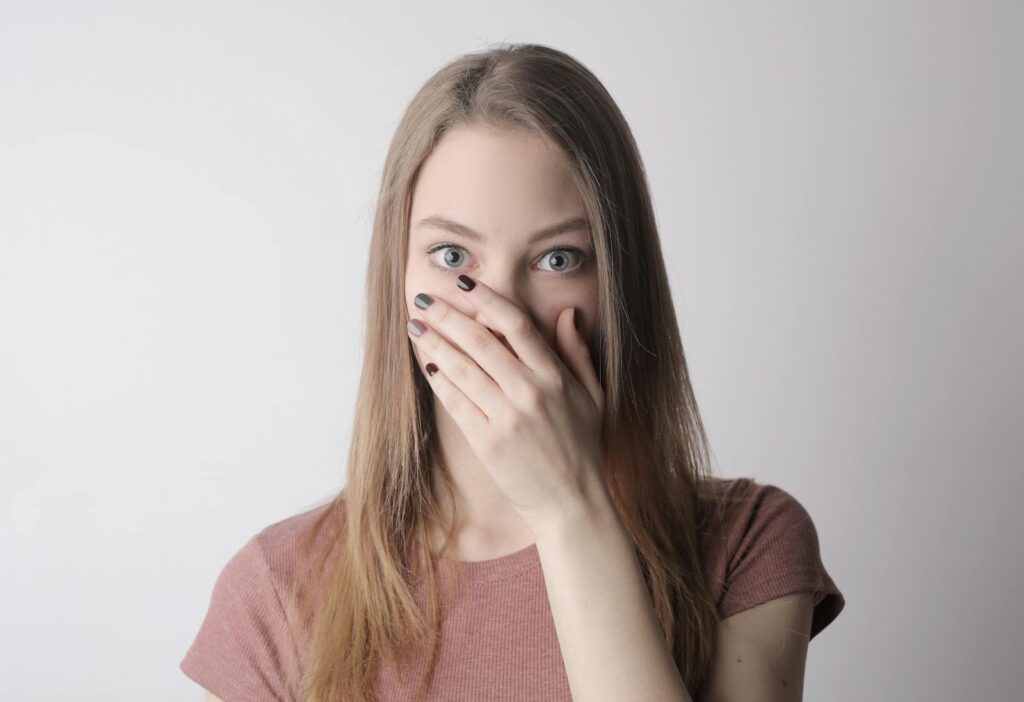 Young woman in pink shirt expressing shock with hand over mouth on simple gray background.