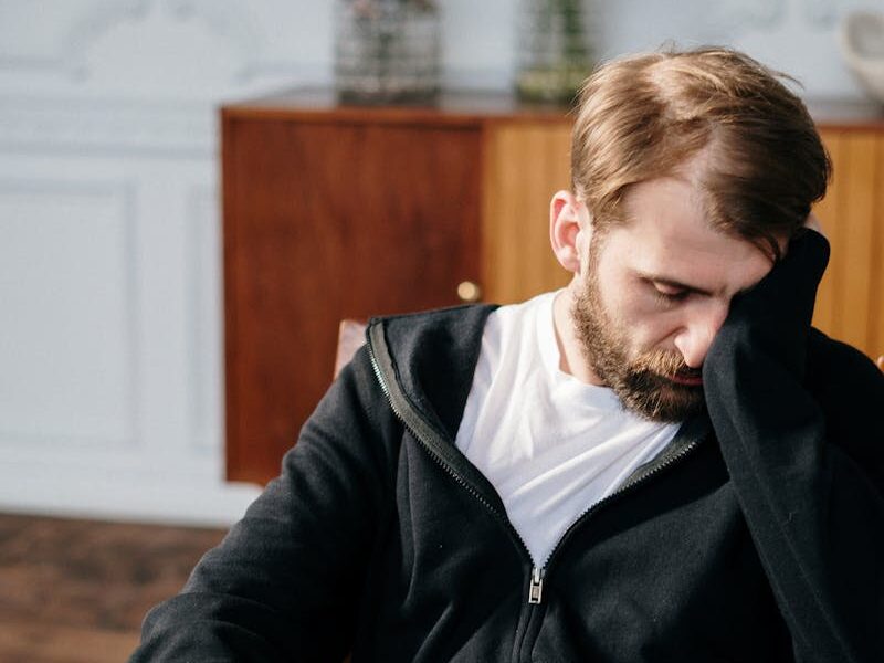 Bearded man in a black hoodie sitting in a chair indoors, appearing deep in thought.