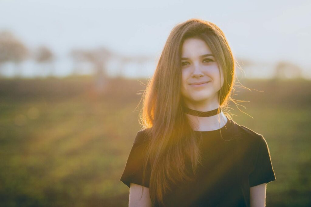 Portrait of a smiling young woman outdoors in a sunlit field, wearing a black choker.