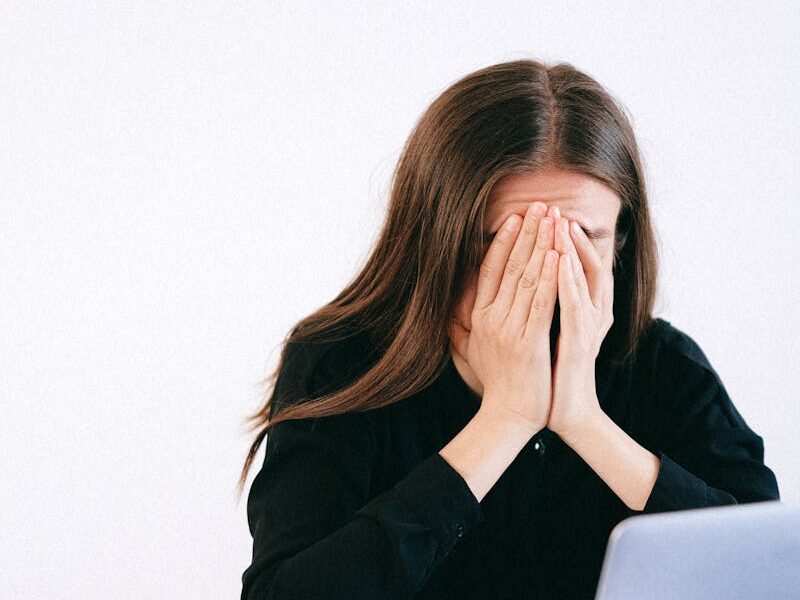 A woman covers her face with hands in stress sitting at an office with a laptop and papers.