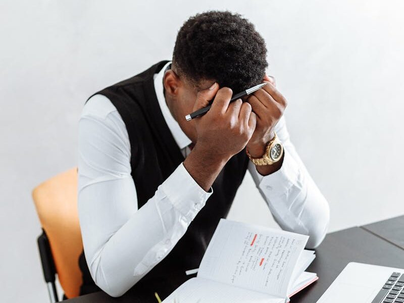 A businessman overwhelmed working indoors with a notebook, laptop, and hourglass.