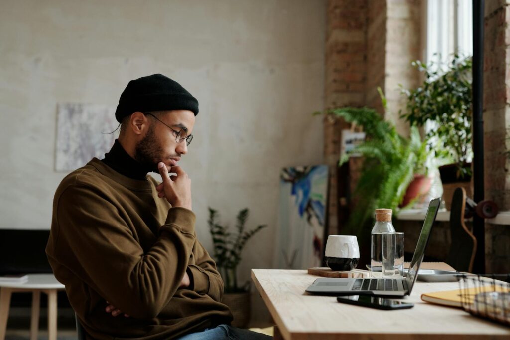 A man in casual attire working thoughtfully on a laptop in a cozy home setting with plants.