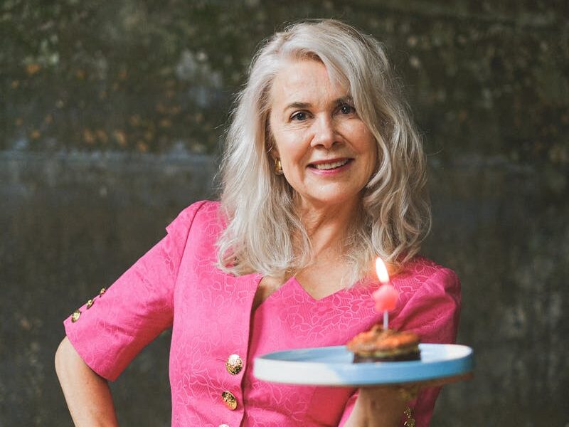 A joyful senior woman in a pink dress carrying a lit birthday cake in an indoor setting.