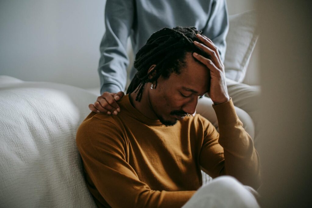 Worried depressed African American male touching head nervously and sitting with eyes closed near supporting wife in bedroom