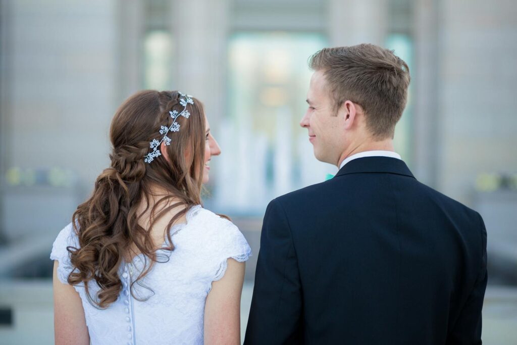 A bride and groom smiling at each other outdoors, dressed in formal wedding attire, exuding joy and elegance.