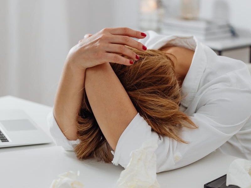 A woman in distress sits at a desk with head in hands, surrounded by tissue papers.