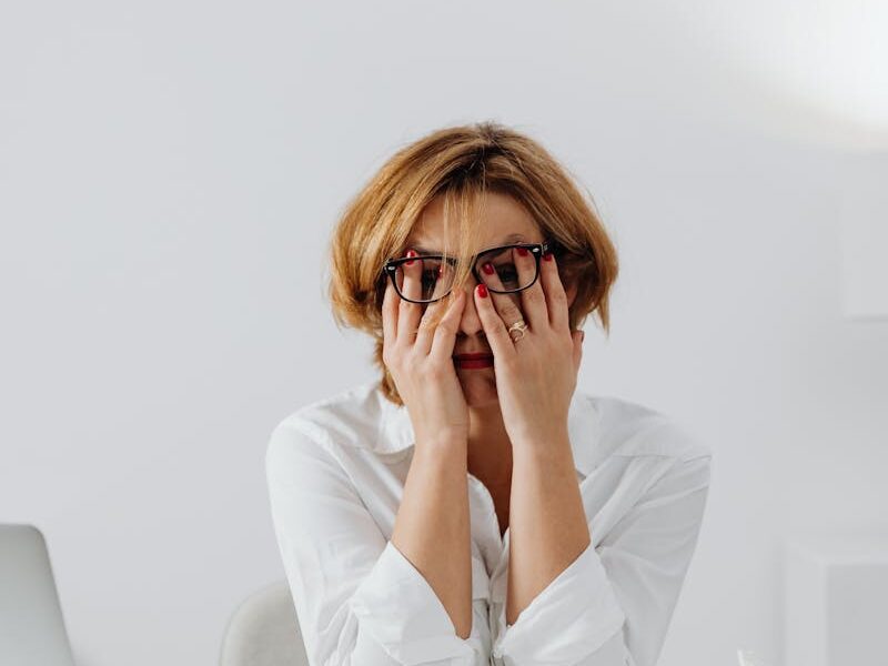 A stressed woman in eyeglasses and white shirt sits at her desk, overwhelmed and exhausted.