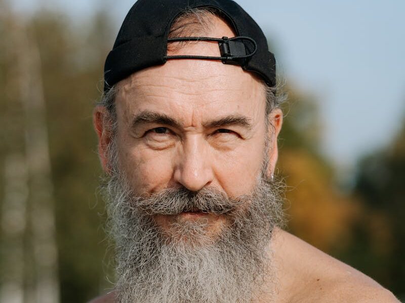 A senior man with a beard and cap poses shirtless outdoors, exuding confidence.