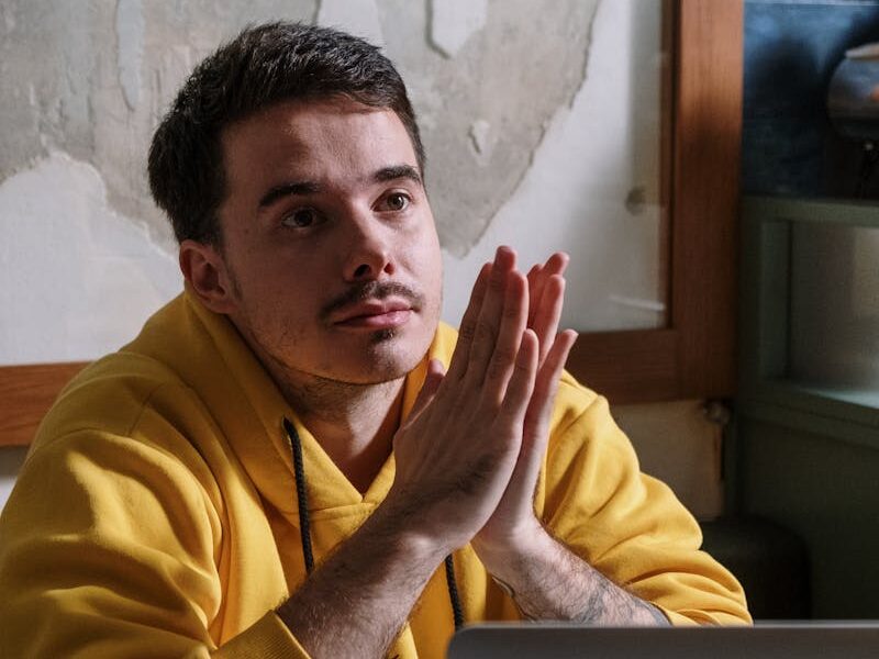 Man in yellow hoodie thinking deeply next to a laptop indoors.
