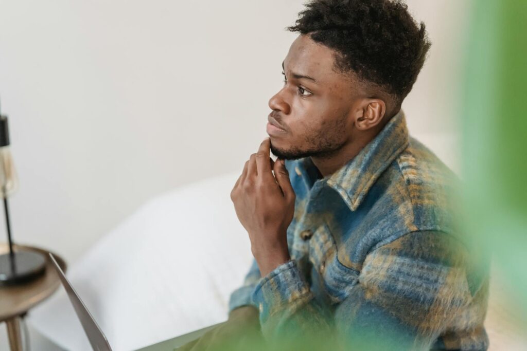 Pensive young man sitting indoors, touching chin, in a peaceful setting, deeply focused.