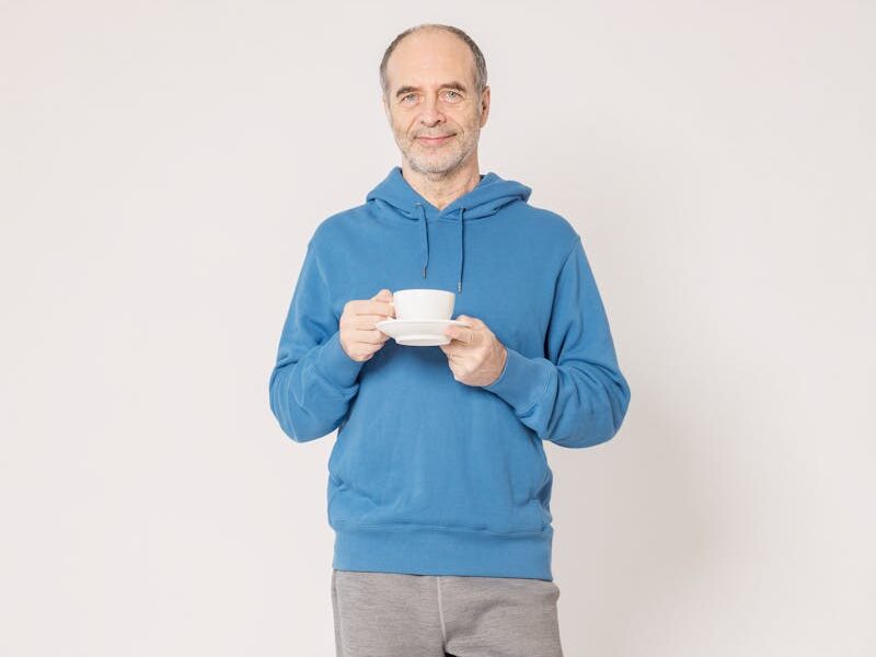 Senior man wearing a blue hoodie, holding a cup indoors, smiling casually.