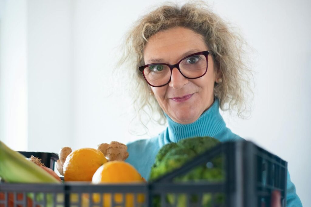 Elderly woman smiling and holding fresh fruit and vegetables indoors.