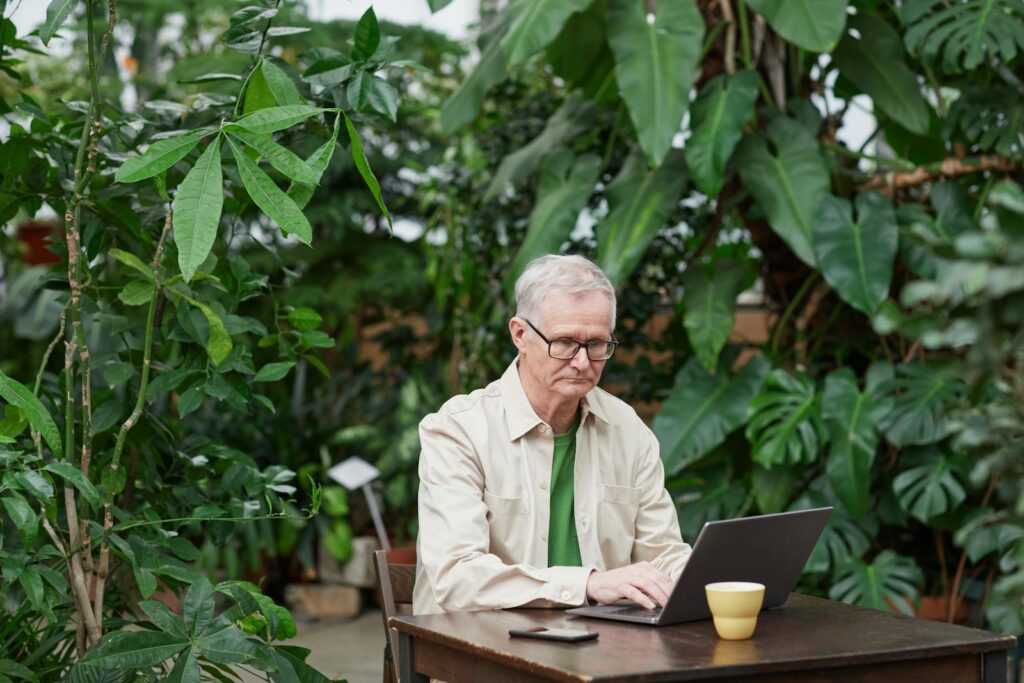 Elder man using laptop in a lush outdoor setting, showcasing freelance remote work lifestyle.