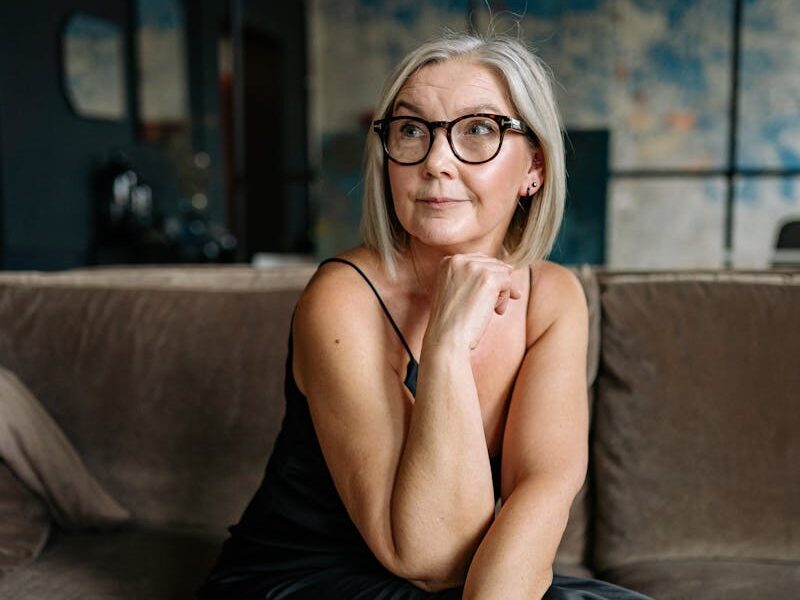 A graceful senior woman in a black dress sitting thoughtfully on a sofa indoors.