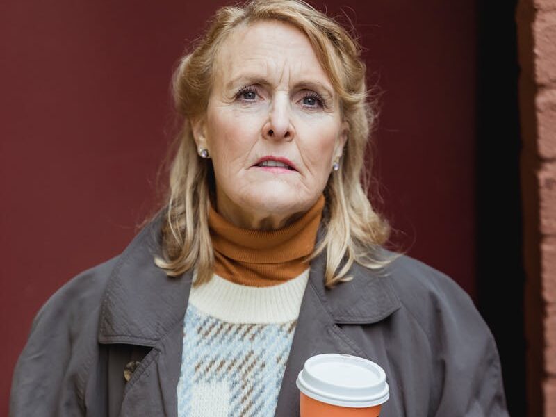 Senior woman in warm clothes holding a coffee cup outdoors on a cool day.