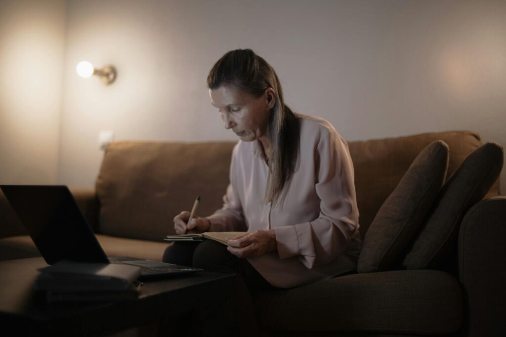 Senior woman sitting on couch, writing notes while using a laptop, cozy indoor setting.