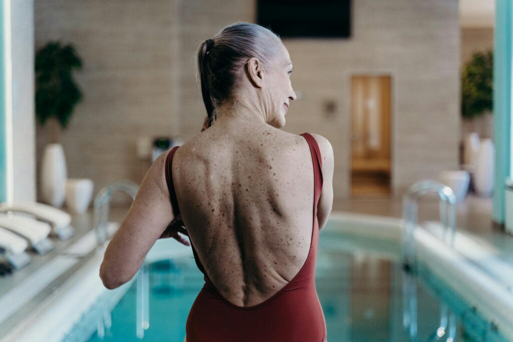 Senior woman in red swimsuit standing at indoor pool side, exuding confidence.