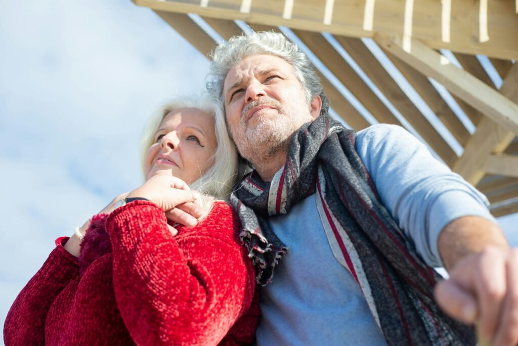 A senior couple enjoys a loving embrace outdoors beneath a wooden pergola, dressed warmly for a cool day.