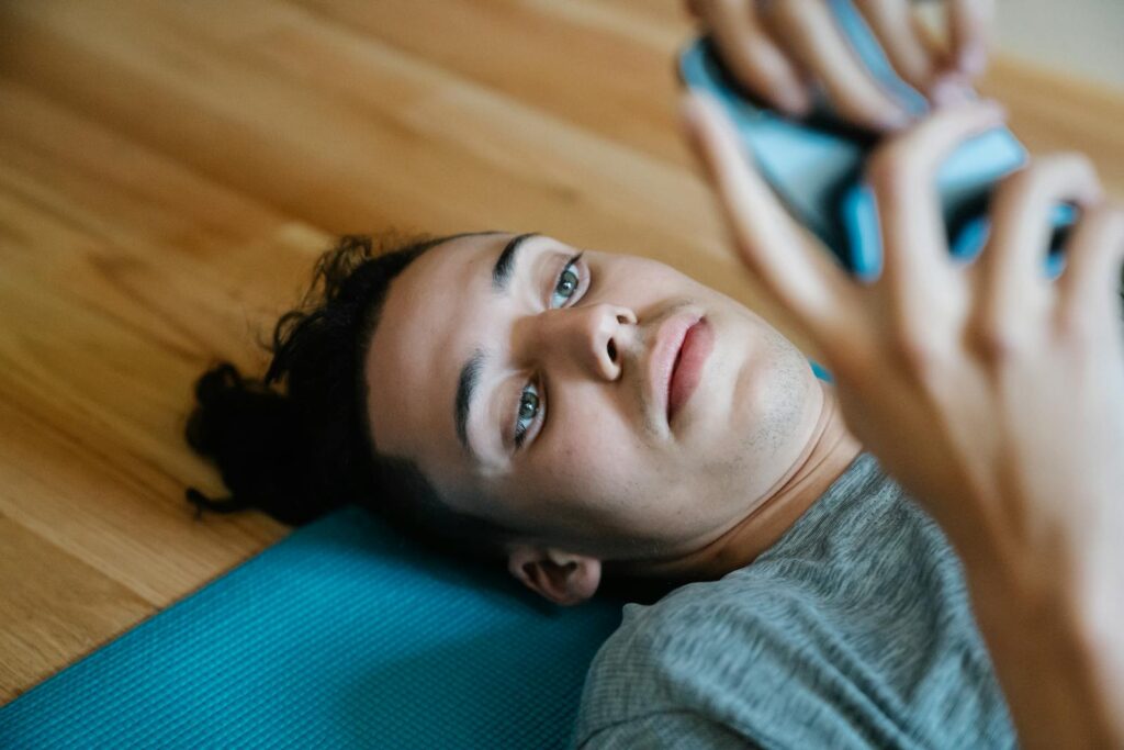 From above of handsome young man with long dark hair using smartphone while lying on blue mat at home