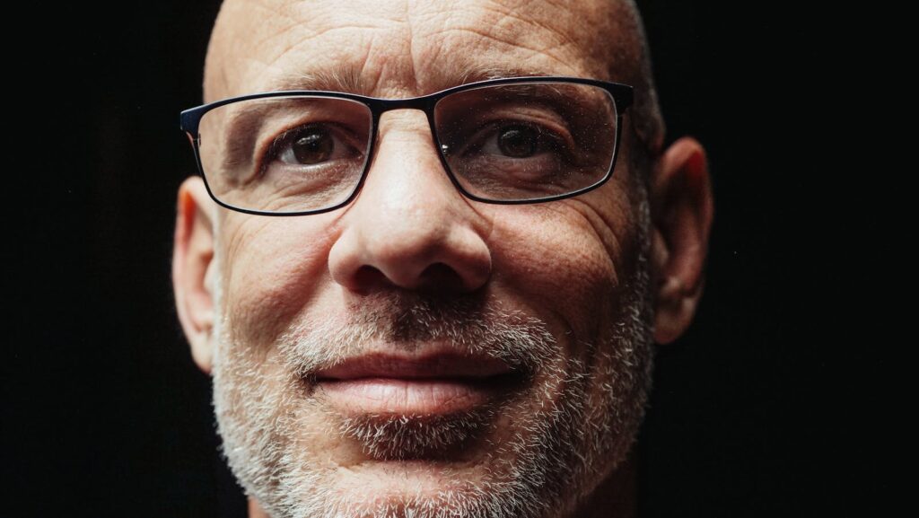 Close-up portrait of a smiling bald elderly man with glasses against a dark background.