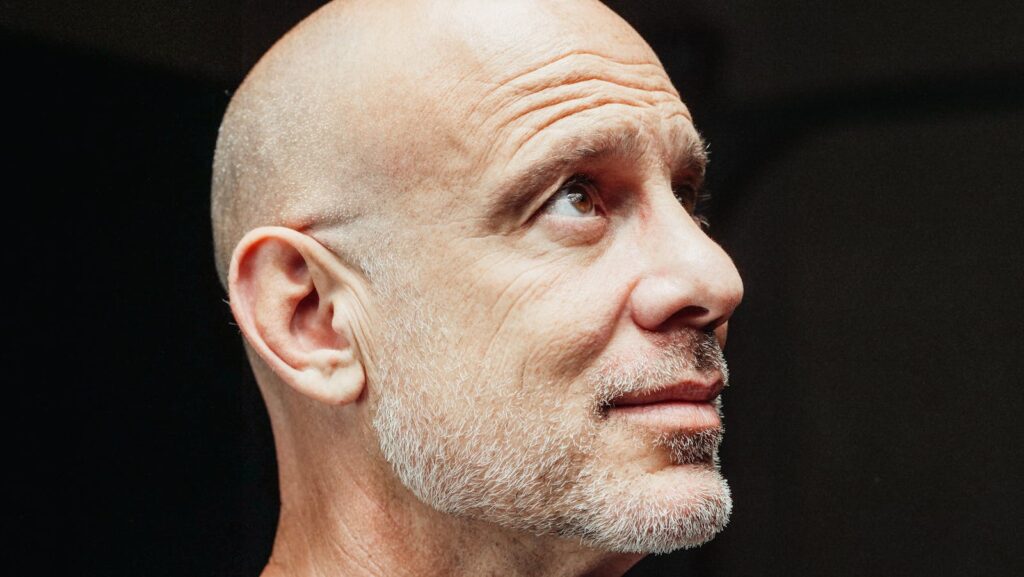 Close-up profile of a senior bald bearded man looking upwards against a dark background.