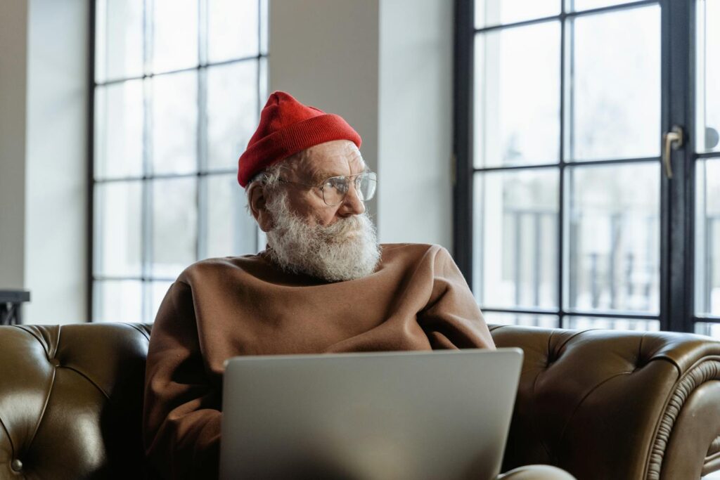 An elderly man in a red beanie is using a laptop indoors, sitting comfortably on a couch.