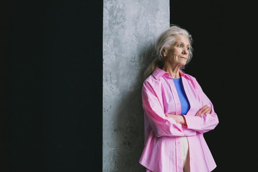 Senior woman with grey hair leaning against a wall, wearing a pink shirt, arms crossed, looking thoughtful.