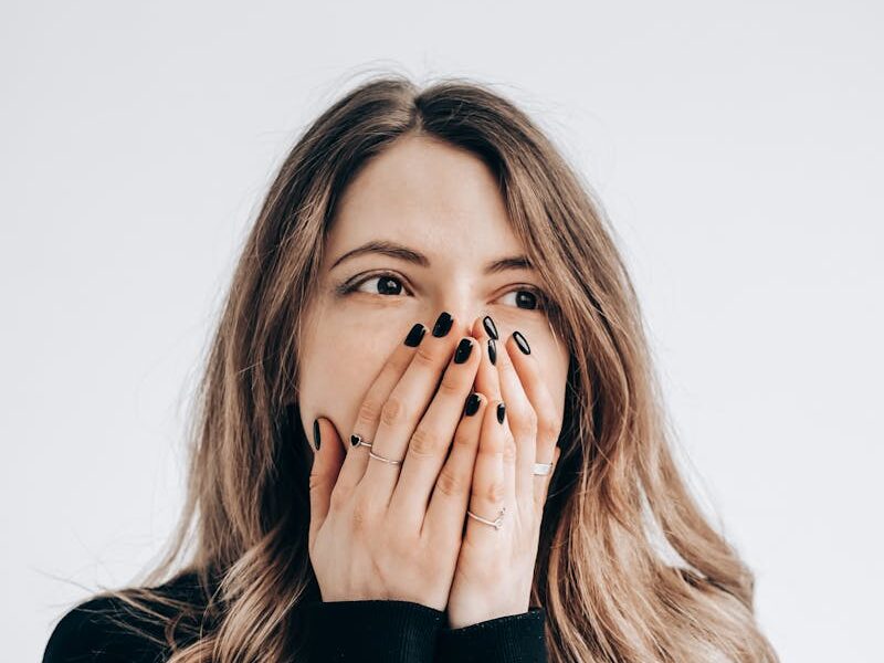 A surprised young woman covers her mouth with her hands, wearing a black sweater.