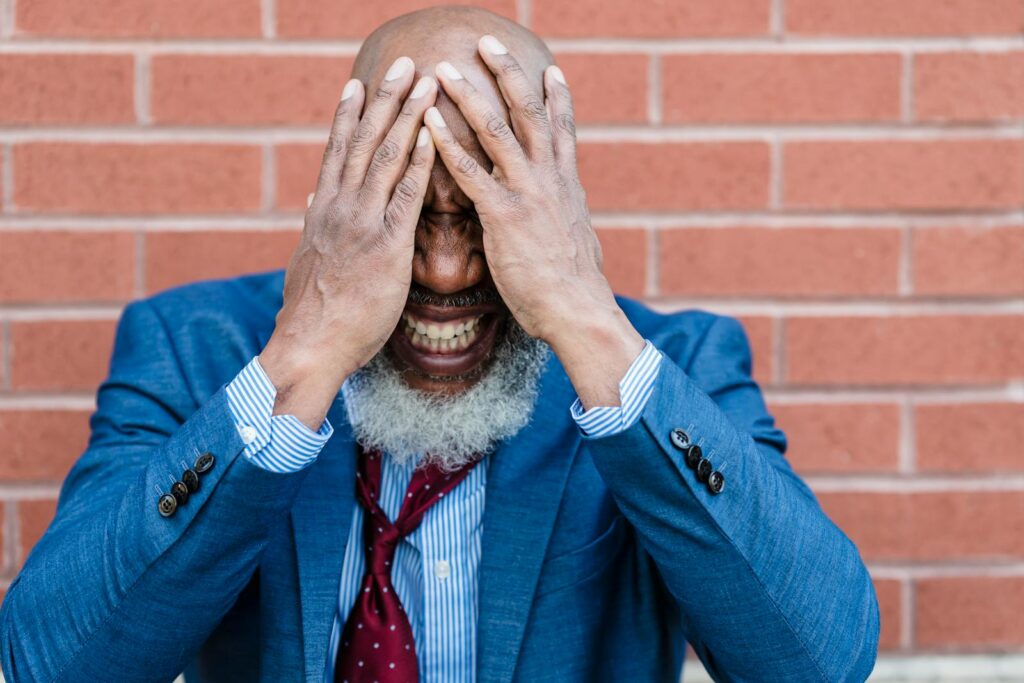 A bearded man in a blue suit looks frustrated, covering his face with his hands in front of a brick wall.