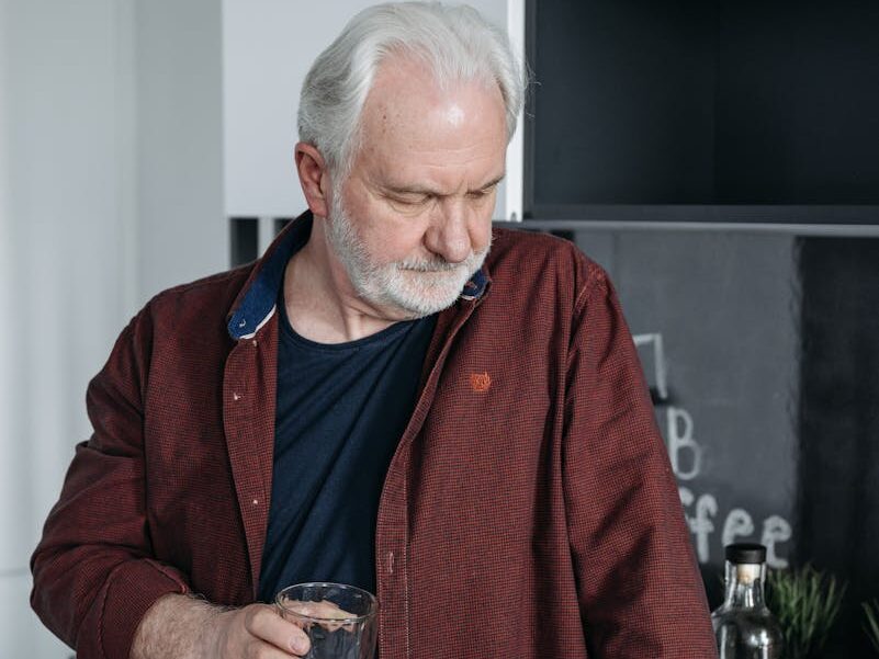 Senior man with gray hair holding a glass, reflecting indoors in a moody atmosphere.