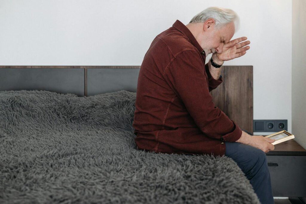 An elderly man sits on a bed in tears holding a picture frame, depicting bereavement.