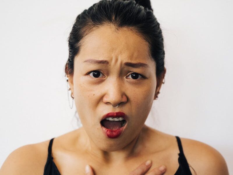 A woman in a black top looks surprised, showcasing a shocked expression with her hand on her chest.