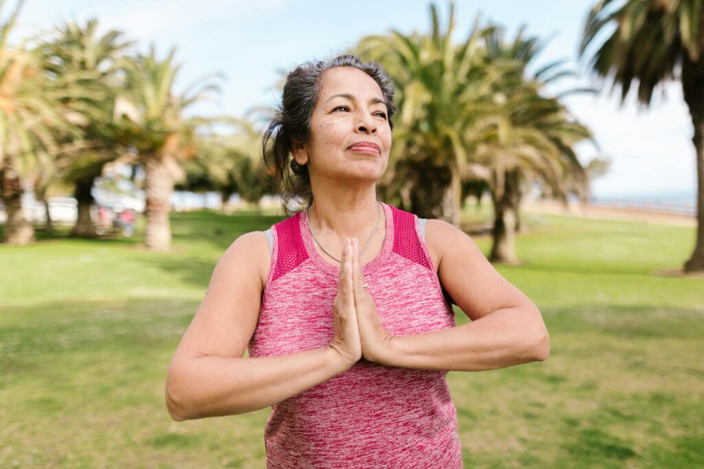 A senior woman in a pink tank top practices yoga in a sunny park surrounded by palm trees.