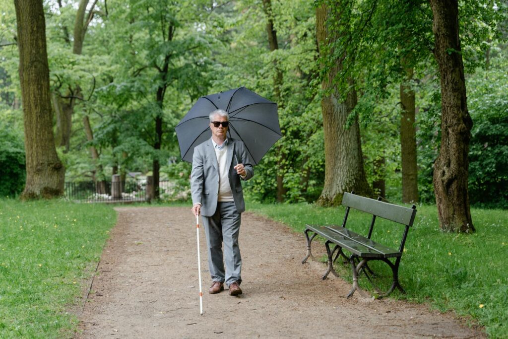 Senior man with visual impairment walks in a park with a walking stick and umbrella.