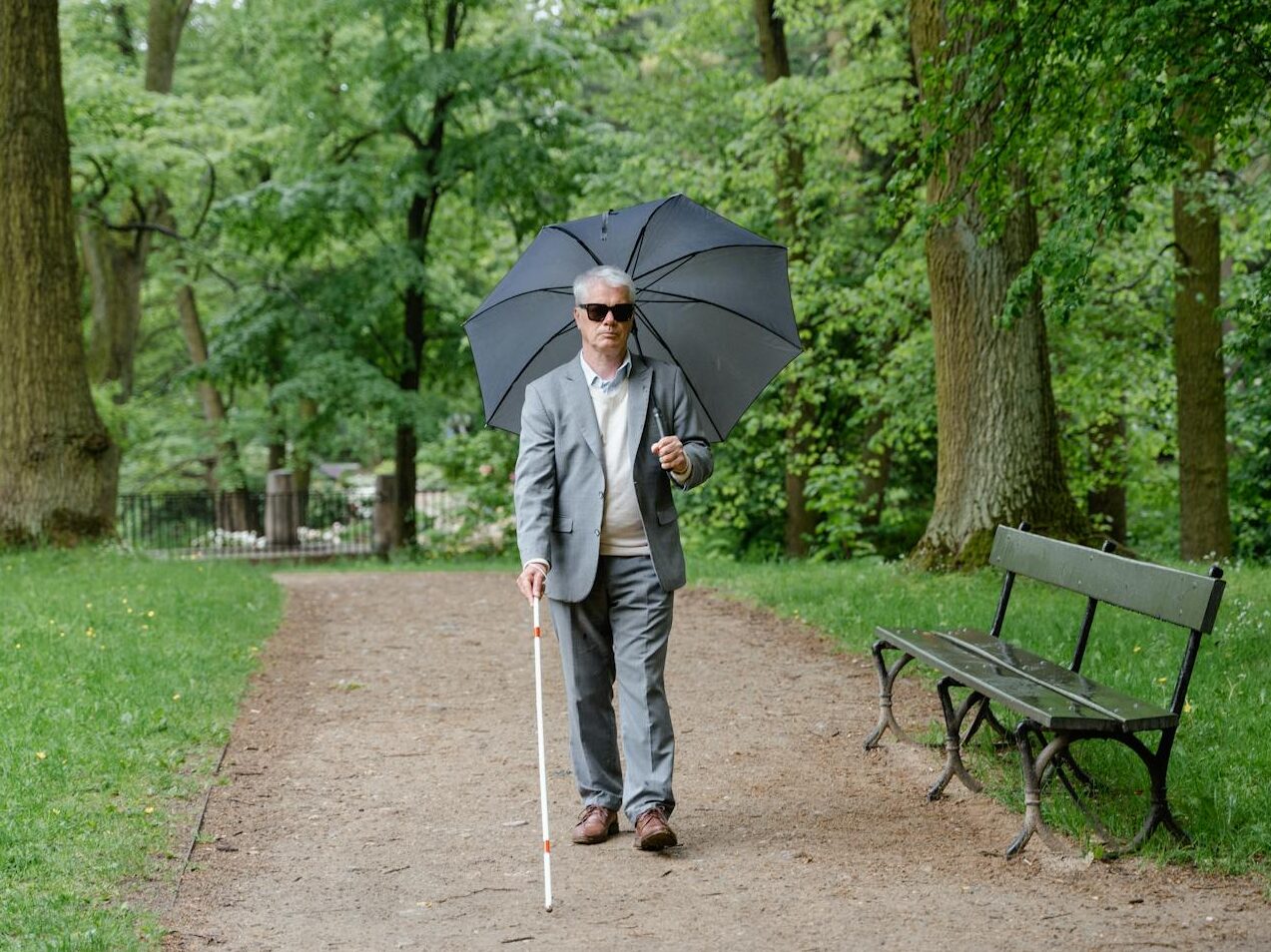 Senior man with visual impairment walks in a park with a walking stick and umbrella.