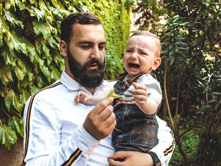 A bearded father comforts his crying baby while holding him in a lush green garden.