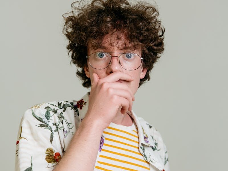 Portrait of a surprised young man wearing glasses and a floral shirt with a striped tee underneath. Neutral background.