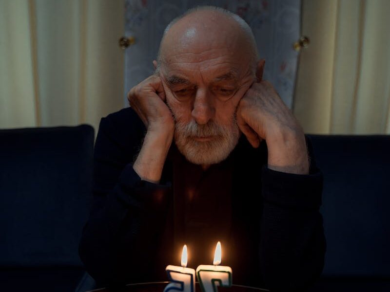 Elderly man sits alone with birthday cake, pondering with lit candles.