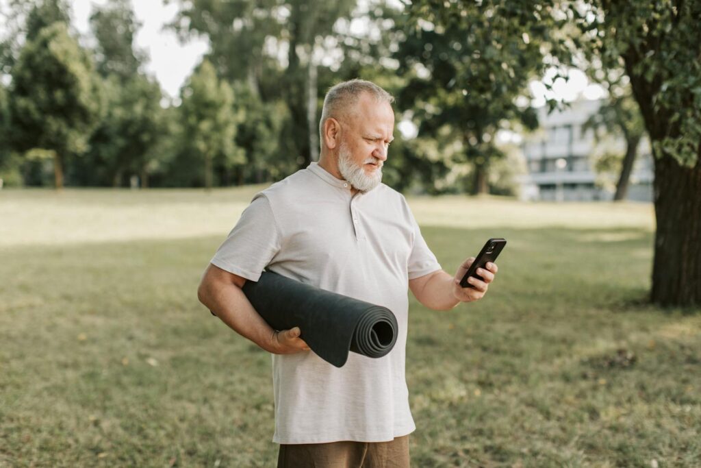 Elderly man with yoga mat texting on phone in a park.