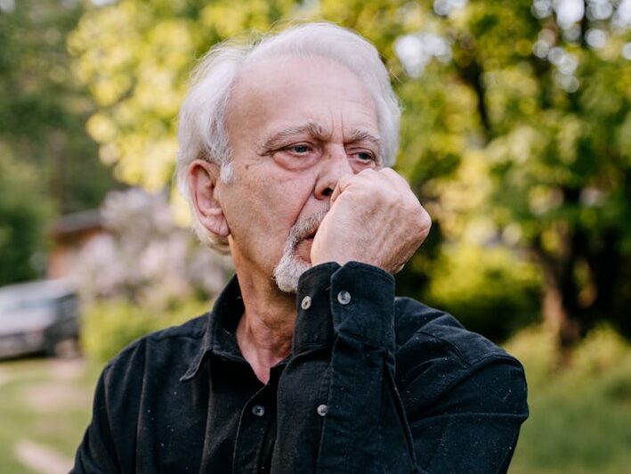 Senior man in a black shirt contemplating while leaning on a walking stick outdoors.