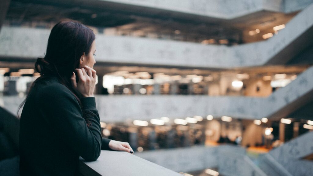 A woman gazes thoughtfully from a balcony in a modern architectural interior space.