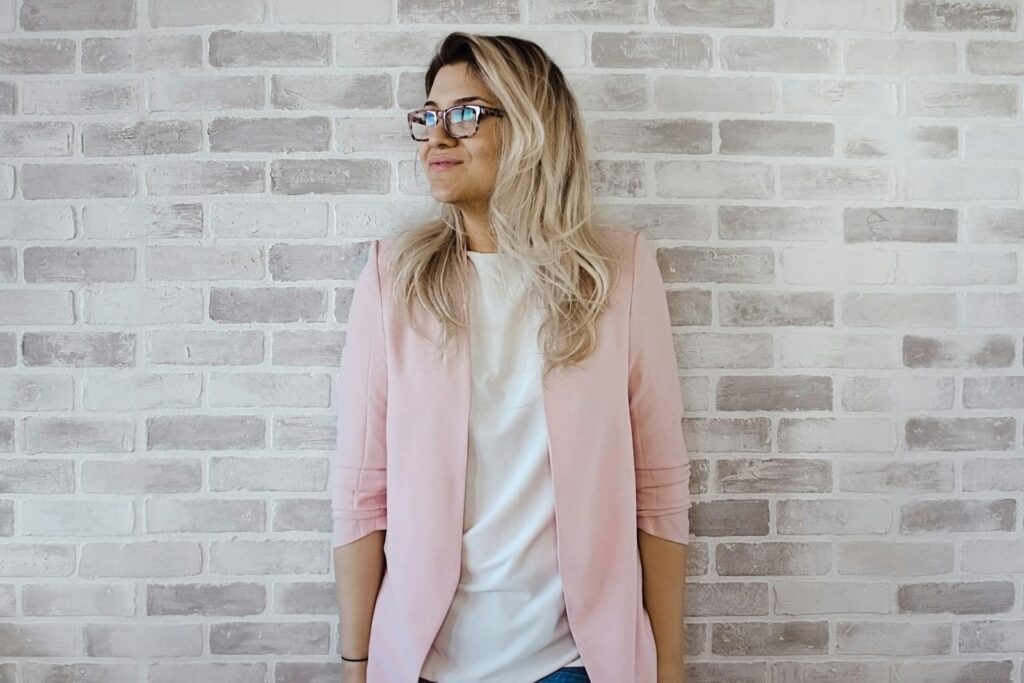 Elegant woman with glasses poses against a brick wall in a trendy pink blazer.