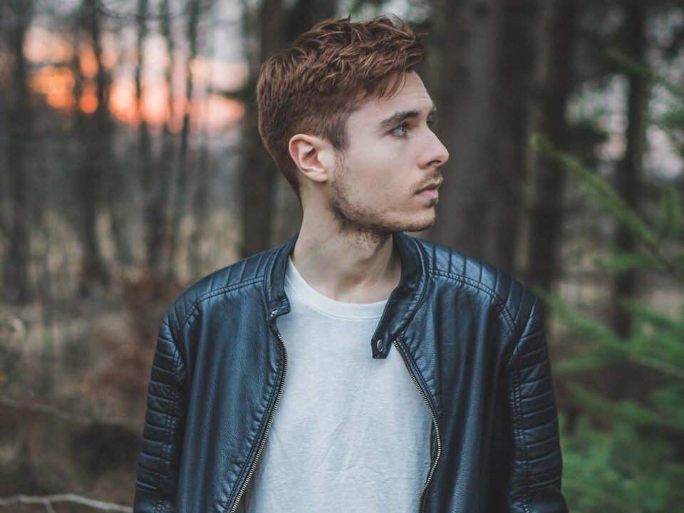 Young man in a black leather jacket standing in a forest during twilight, styled fashion portrait.