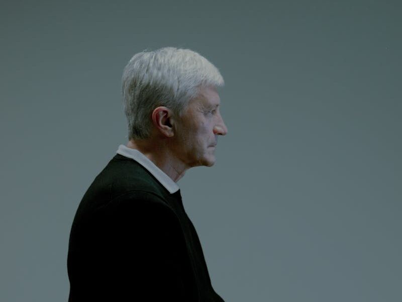 Portrait of a senior man in profile sitting indoors on a chair with grey background.