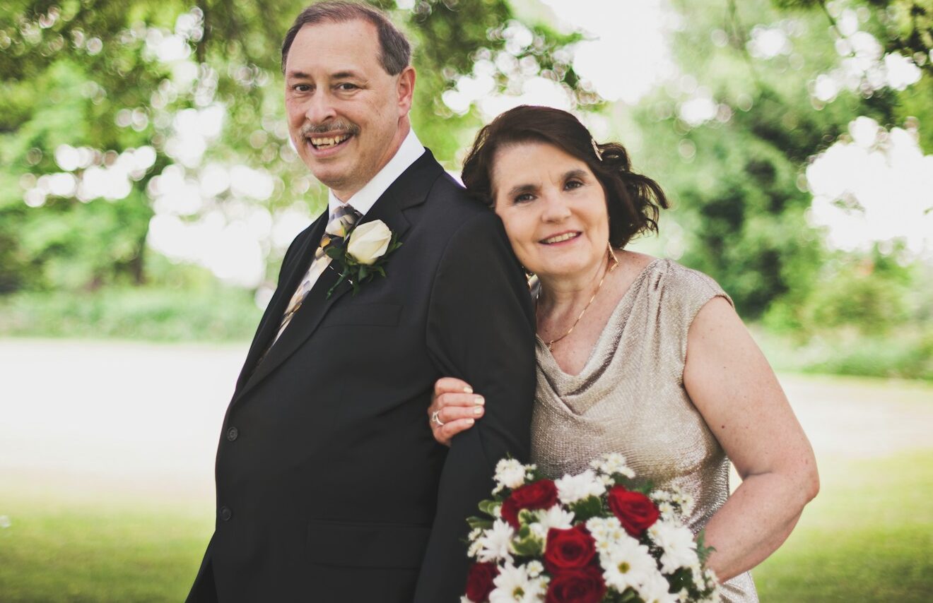 man in black suit standing beside woman in gray sleeveless dress holding bouquet of flowers