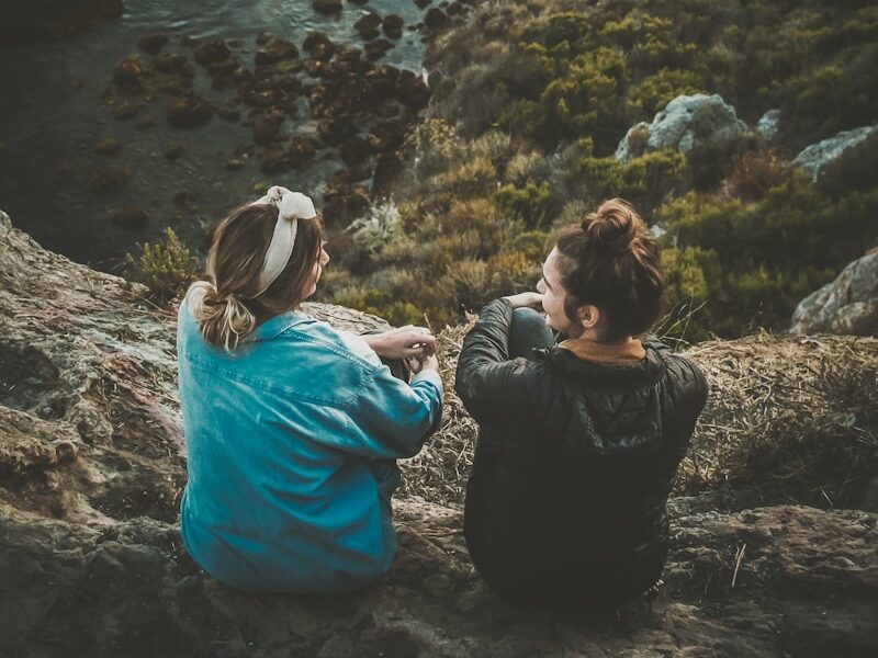 two women sitting on cliff while chatting viewing blue body of water