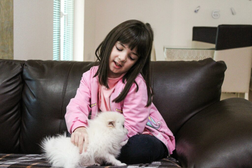 a little girl sitting on a couch petting a small white dog